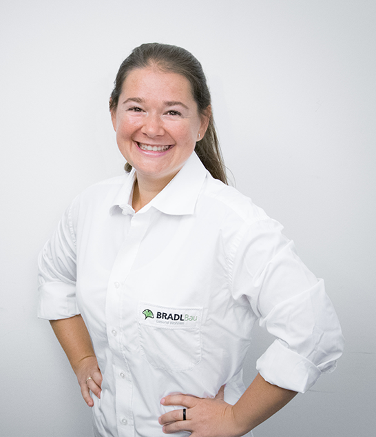 Alexandra Hochreiter, lächelnd, in einem weißen Hemd mit BRADL Bau-Logo, die selbstbewusst vor einem weißen Hintergrund posiert.