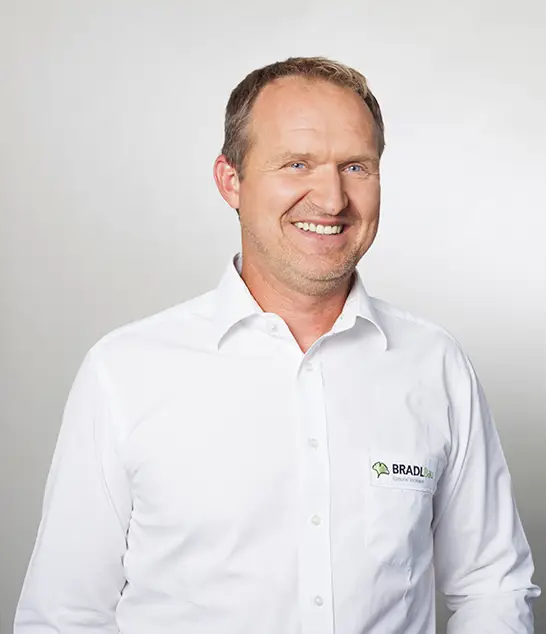 Armin Höfl, lächelnd im weißen "BRADLBAU"-Hemd, Oberbauleiter, steht vor einem schlichten Hintergrund.