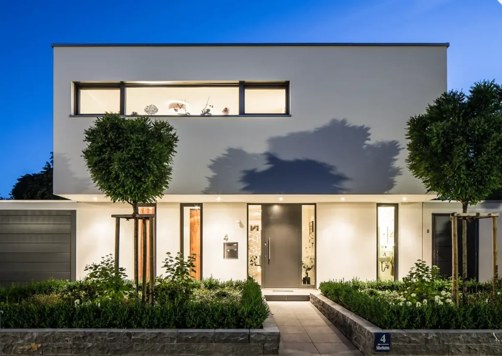 Modernes Haus mit Glasfassade, großen Fenstern und gepflegtem Grün, stimmungsvoll bei Dämmerung beleuchtet.