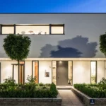 Modernes Haus mit Glasfassade, großen Fenstern und gepflegtem Grün, stimmungsvoll bei Dämmerung beleuchtet.