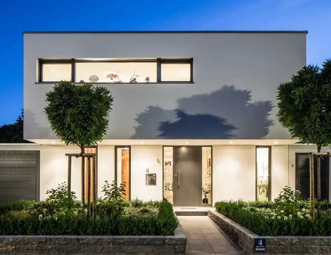 Modernes Haus mit Glasfassade, großen Fenstern und gepflegtem Grün, stimmungsvoll bei Dämmerung beleuchtet.