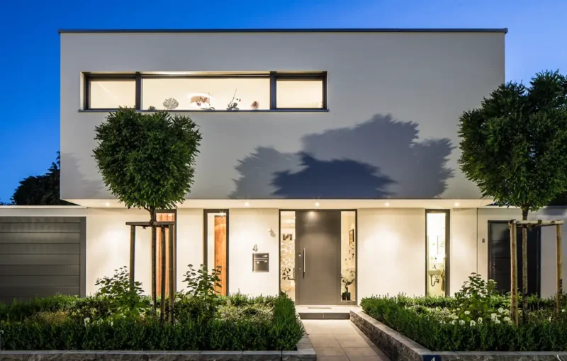 Modernes Haus mit Glasfassade, großen Fenstern und gepflegtem Grün, stimmungsvoll bei Dämmerung beleuchtet.