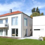 Modernes zweistöckiges weißes Haus mit großen Fenstern, rotem Dach, Balkon und grünem Rasen unter blauem Himmel.