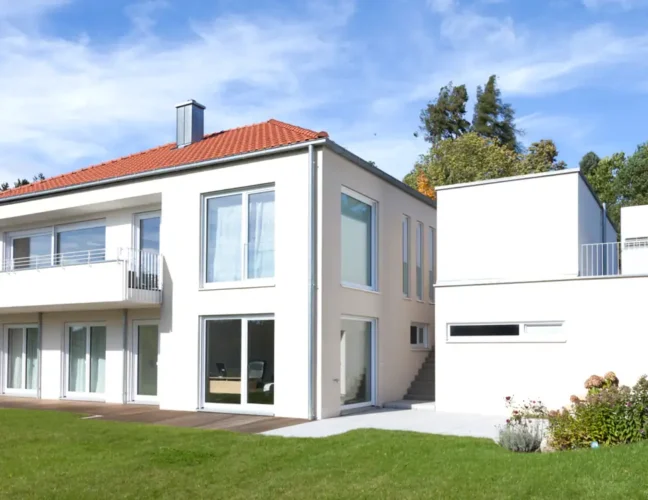 Modernes zweistöckiges weißes Haus mit großen Fenstern, rotem Dach, Balkon und grünem Rasen unter blauem Himmel.