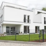 Modernes weißes zweistöckiges Haus mit großen Fenstern, einem eingezäunten Hof und einem kleinen Vorgarten.