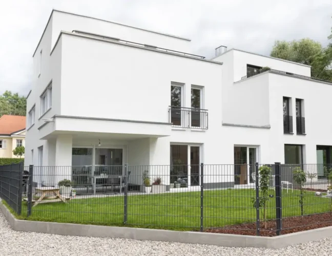 Modernes weißes zweistöckiges Haus mit großen Fenstern, einem eingezäunten Hof und einem kleinen Vorgarten.