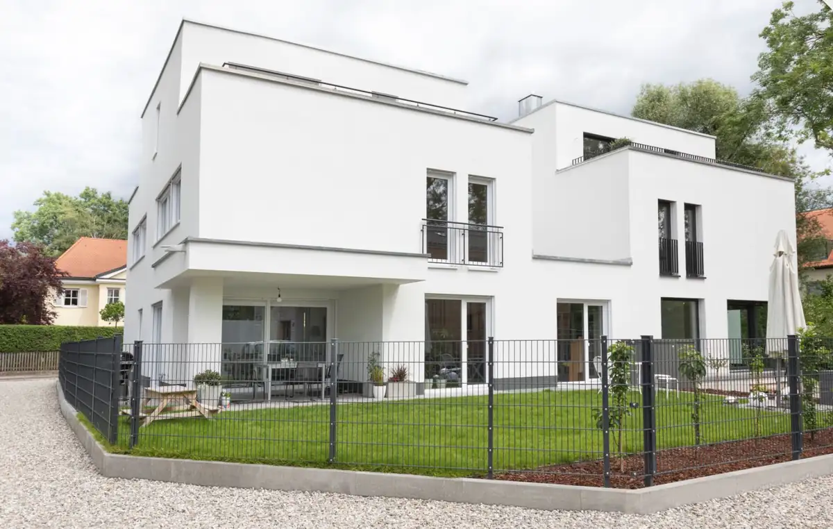 Modernes weißes zweistöckiges Haus mit großen Fenstern, einem eingezäunten Hof und einem kleinen Vorgarten.