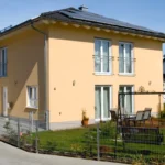 Modernes zweistöckiges gelbes Haus mit kleinem Garten, Terrassenmöbeln und einer Auffahrt an einem sonnigen Tag.