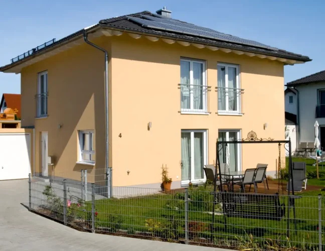 Modernes zweistöckiges gelbes Haus mit kleinem Garten, Terrassenmöbeln und einer Auffahrt an einem sonnigen Tag.