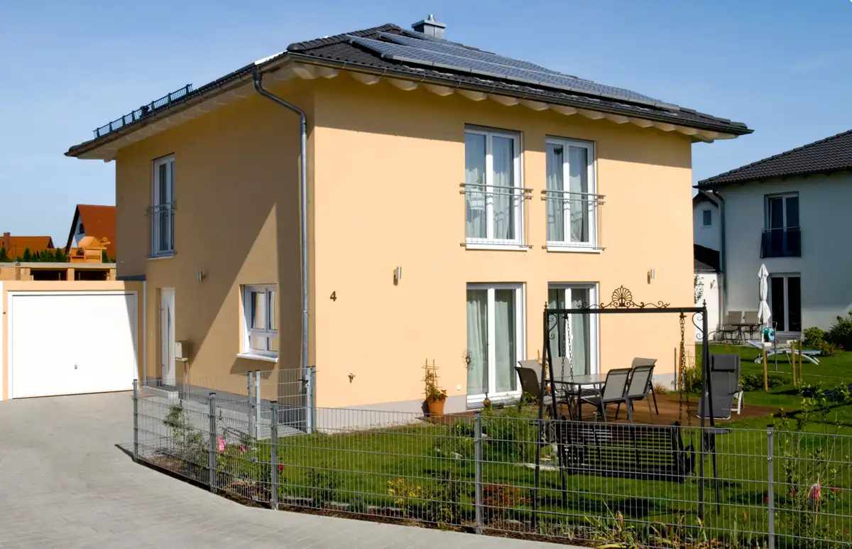 Modernes zweistöckiges gelbes Haus mit kleinem Garten, Terrassenmöbeln und einer Auffahrt an einem sonnigen Tag.