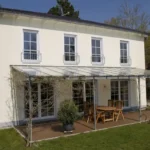 Zweistöckiges weißes Haus mit hohen Fenstern, Terrassenmöbeln und einer Pergola in einem grünen Garten.