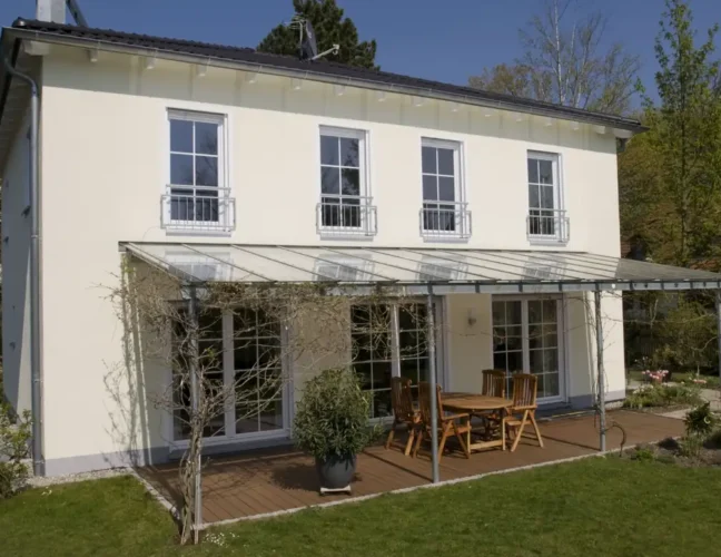 Zweistöckiges weißes Haus mit hohen Fenstern, Terrassenmöbeln und einer Pergola in einem grünen Garten.