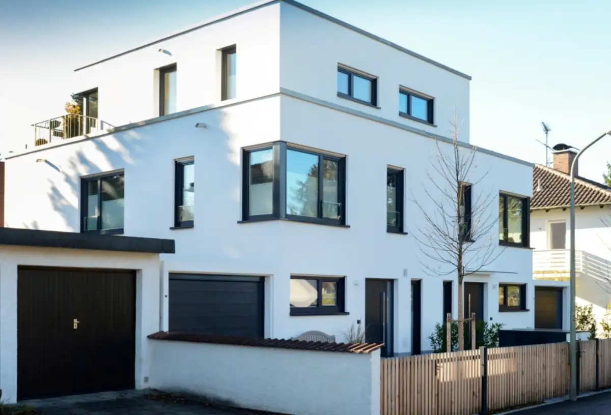 Modernes weißes zweistöckiges Haus mit Flachdach, großen Fenstern, Garage und kleinem eingezäunten Hof an einem sonnigen Tag.