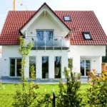 Modernes Haus mit rotem Dach, weißen Wänden, großen Fenstern und einem grünen Garten vor dem Haus.