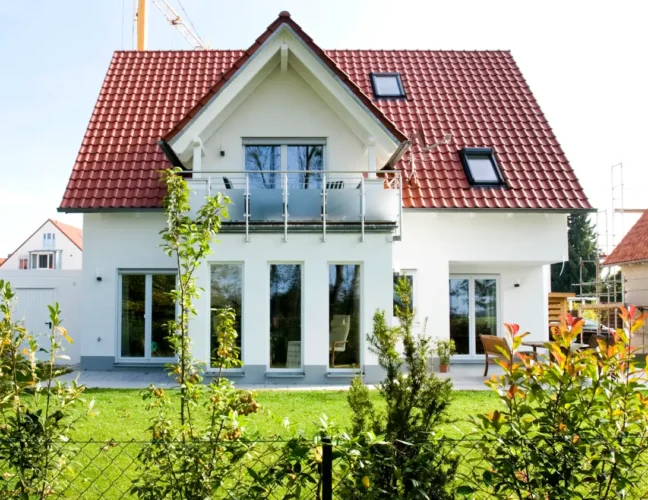 Modernes Haus mit rotem Dach, weißen Wänden, großen Fenstern und einem grünen Garten vor dem Haus.