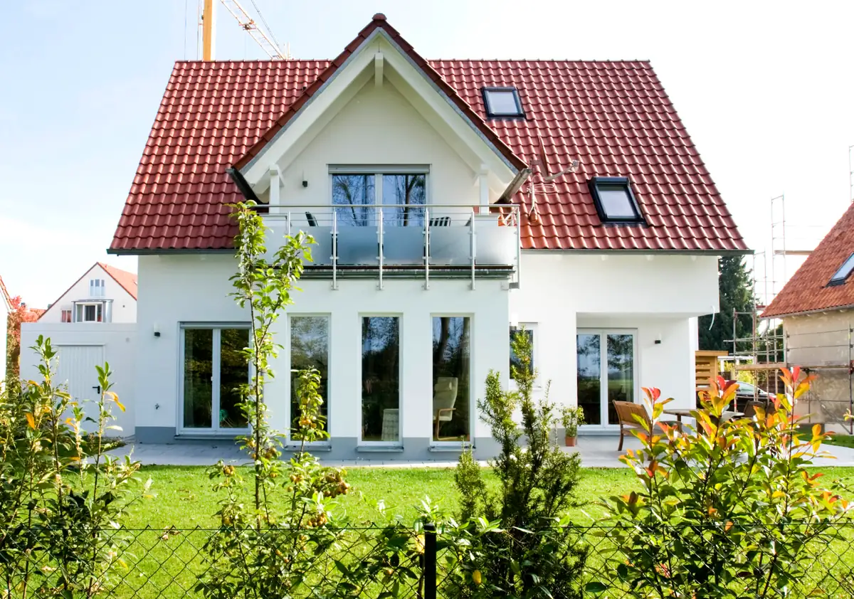 Modernes Haus mit rotem Dach, weißen Wänden, großen Fenstern und einem grünen Garten vor dem Haus.
