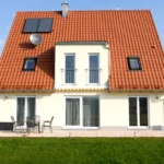 Ein modernes Haus mit rotem Ziegeldach, Sonnenkollektoren und einer Veranda in einem grasbewachsenen Hof unter blauem Himmel.