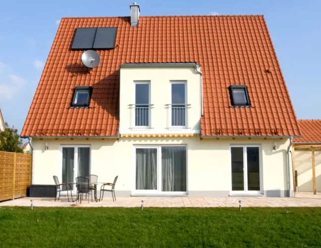 Ein modernes Haus mit rotem Ziegeldach, Sonnenkollektoren und einer Veranda in einem grasbewachsenen Hof unter blauem Himmel.