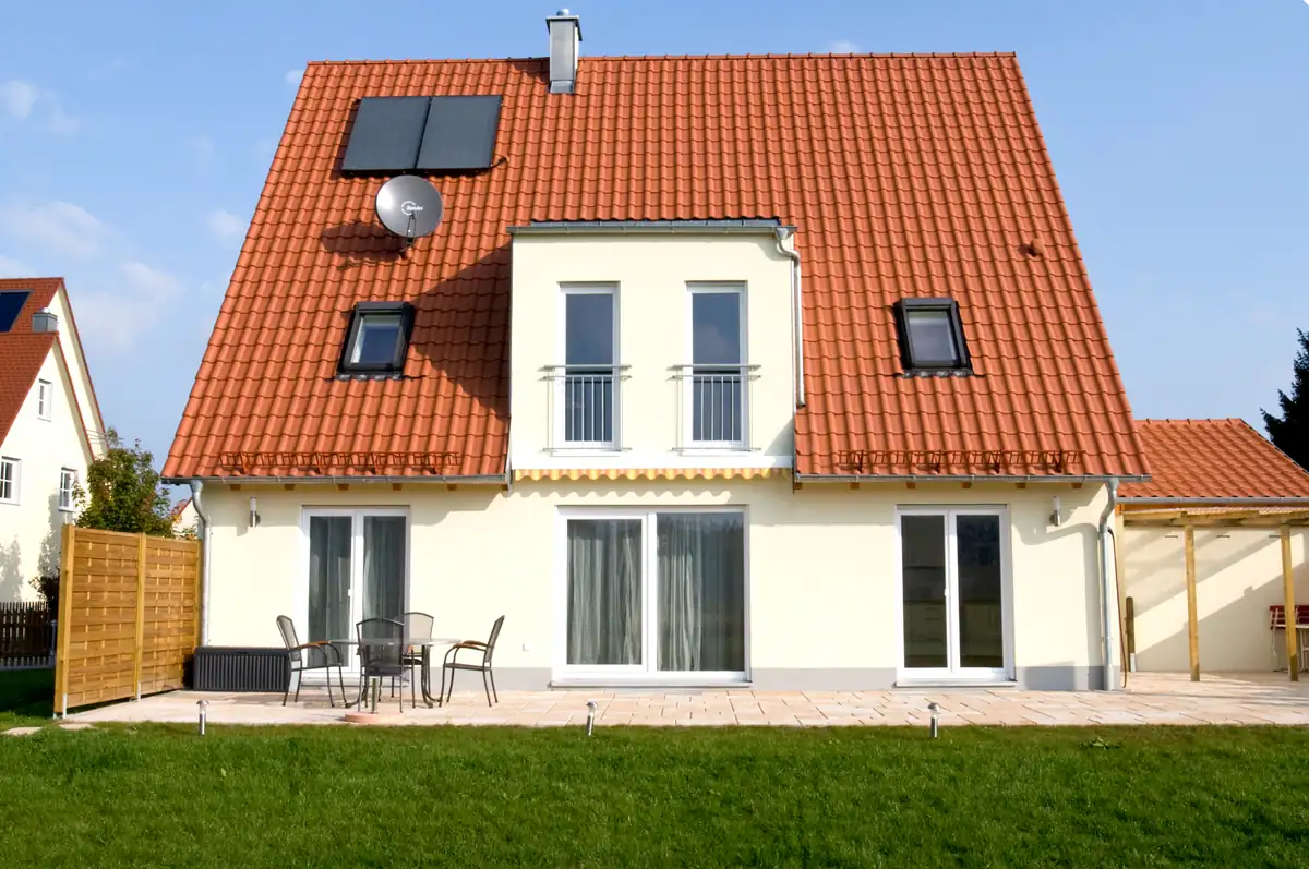 Ein modernes Haus mit rotem Ziegeldach, Sonnenkollektoren und einer Veranda in einem grasbewachsenen Hof unter blauem Himmel.