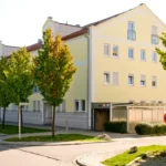 Ein gelbes Wohnhaus an einem sonnigen Tag, mit Bäumen und geparkten Autos entlang der Straße.