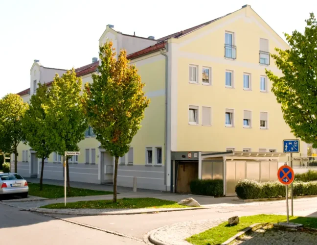 Ein gelbes Wohnhaus an einem sonnigen Tag, mit Bäumen und geparkten Autos entlang der Straße.