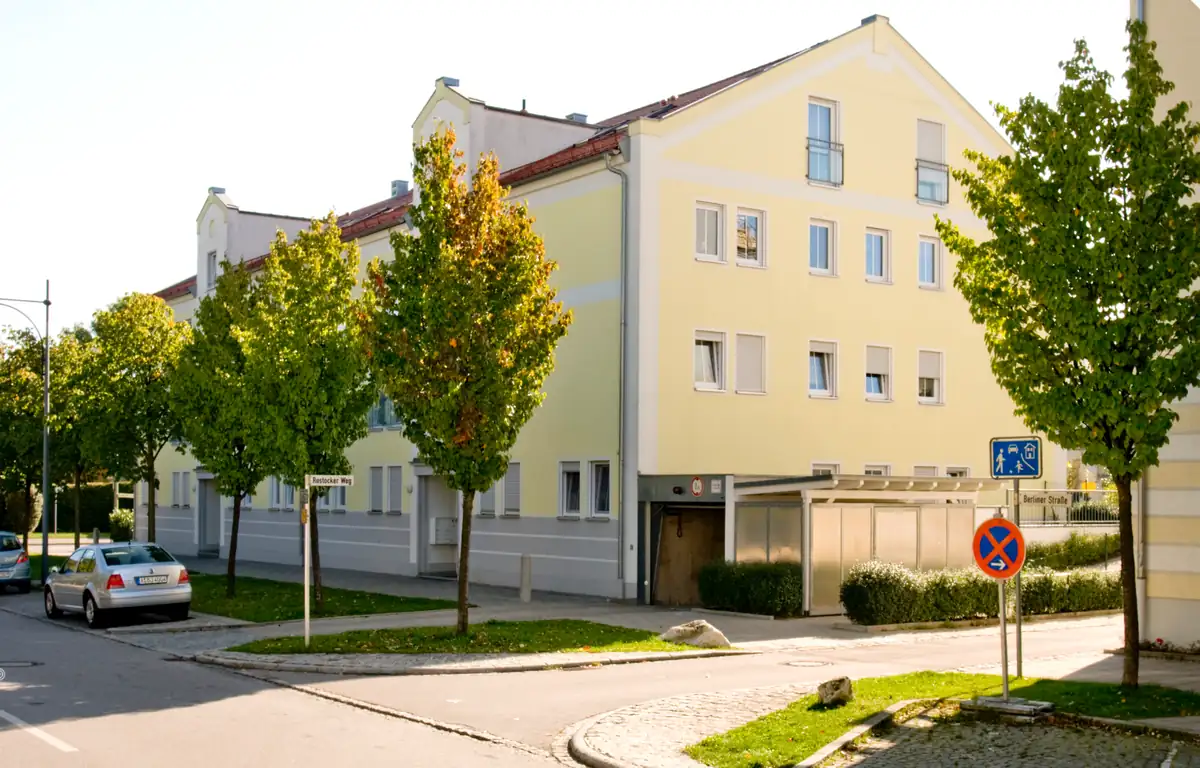 Ein gelbes Wohnhaus an einem sonnigen Tag, mit Bäumen und geparkten Autos entlang der Straße.