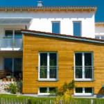 Modernes Haus mit weißen Wänden und einem Holzanbau, großen Fenstern und einem kleinen Vorgarten.