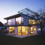 Modernes zweistöckiges Haus mit großen, in der Dämmerung beleuchteten Fenstern, mit Glasbalkon und -terrasse.