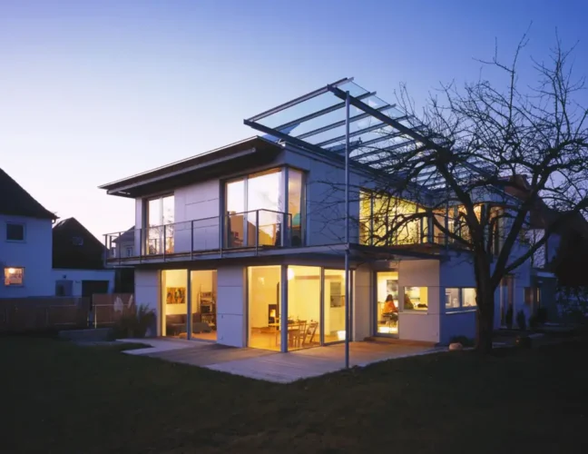 Modernes zweistöckiges Haus mit großen, in der Dämmerung beleuchteten Fenstern, mit Glasbalkon und -terrasse.