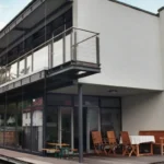 Modernes zweistöckiges Haus mit großen Fenstern, Balkon und Sitzgelegenheit im Freien auf einer Holzterrasse.