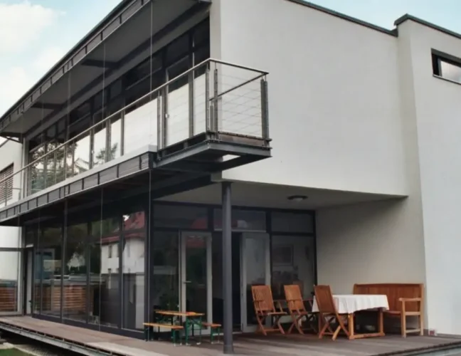 Modernes zweistöckiges Haus mit großen Fenstern, Balkon und Sitzgelegenheit im Freien auf einer Holzterrasse.