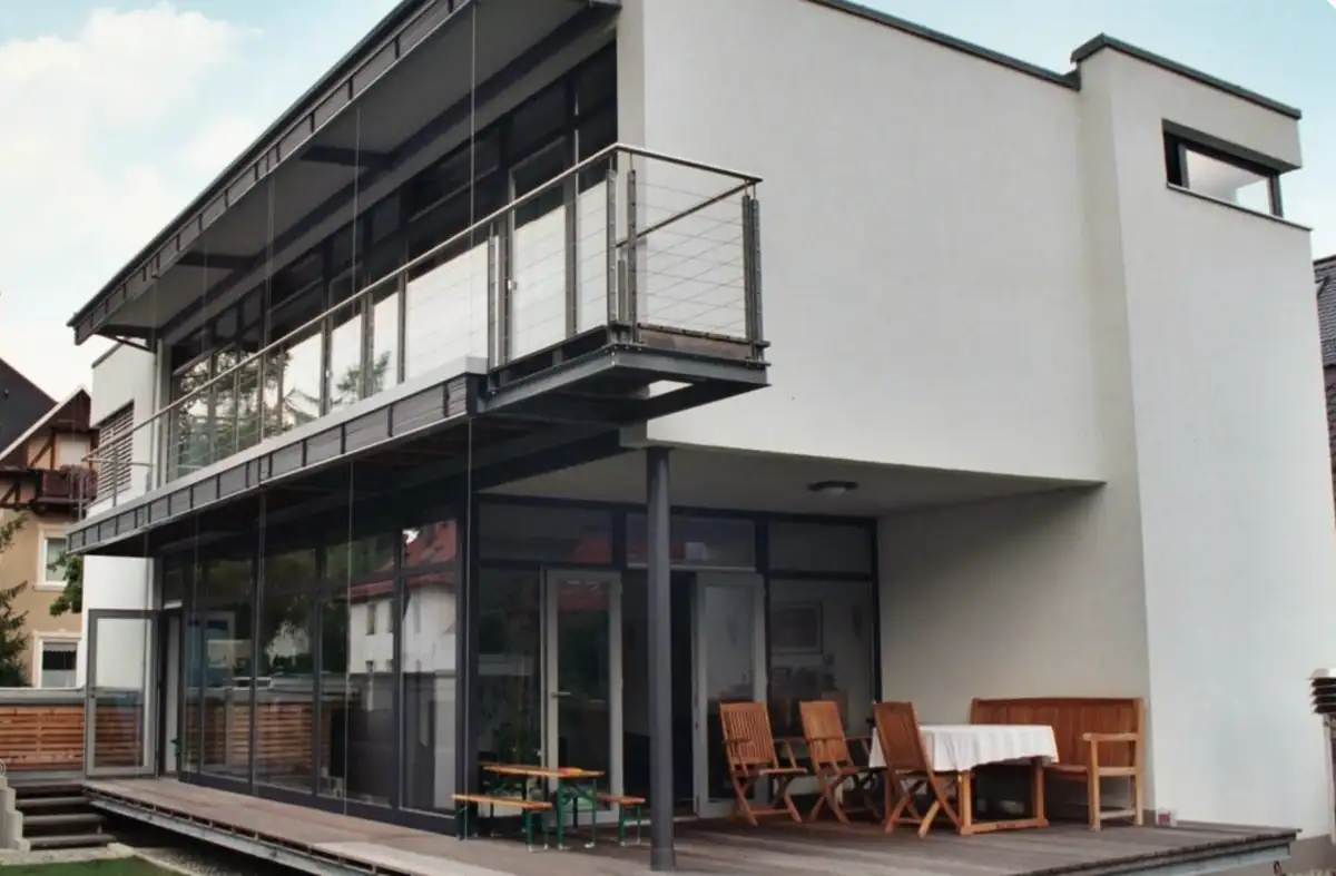 Modernes zweistöckiges Haus mit großen Fenstern, Balkon und Sitzgelegenheit im Freien auf einer Holzterrasse.