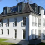 Modernes weißes dreistöckiges Haus mit großen Fenstern und Sonnenkollektoren auf dem Dach an einem sonnigen Tag.