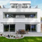 Modernes, weißes, zweistöckiges Haus mit großen Fenstern, einer Terrasse und einem Essplatz im Freien im Hinterhof.