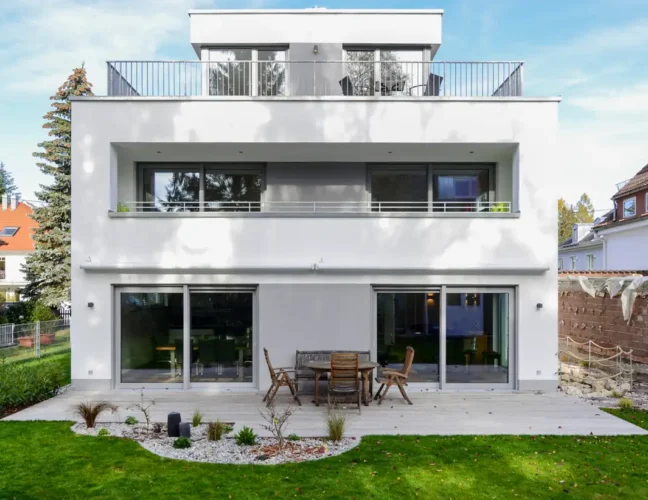 Modernes, weißes, zweistöckiges Haus mit großen Fenstern, einer Terrasse und einem Essplatz im Freien im Hinterhof.