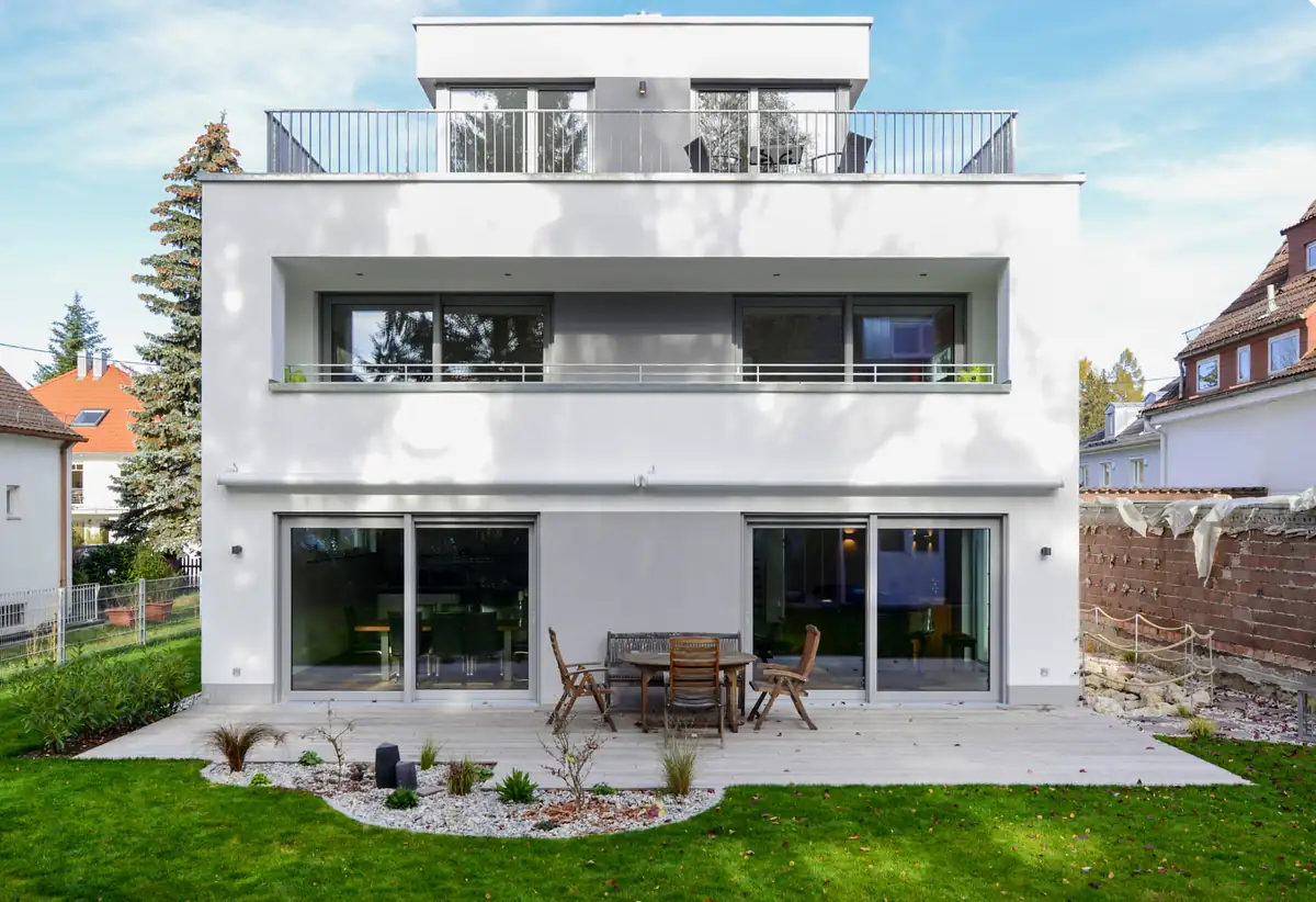Modernes, weißes, zweistöckiges Haus mit großen Fenstern, einer Terrasse und einem Essplatz im Freien im Hinterhof.