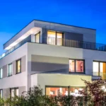 Modernes dreistöckiges Haus mit großen Fenstern, Flachdach und Außenbeleuchtung in der Abenddämmerung.