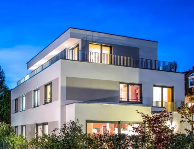 Modernes dreistöckiges Haus mit großen Fenstern, Flachdach und Außenbeleuchtung in der Abenddämmerung.