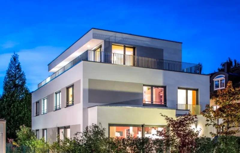 Modernes dreistöckiges Haus mit großen Fenstern, Flachdach und Außenbeleuchtung in der Abenddämmerung.