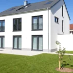 Modernes zweistöckiges weißes Haus mit großen Fenstern, einer Garage und einem kleinen Baum im grasbewachsenen Hof.