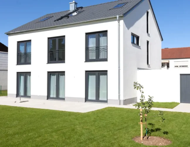 Modernes zweistöckiges weißes Haus mit großen Fenstern, einer Garage und einem kleinen Baum im grasbewachsenen Hof.