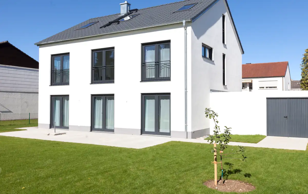 Modernes zweistöckiges weißes Haus mit großen Fenstern, einer Garage und einem kleinen Baum im grasbewachsenen Hof.