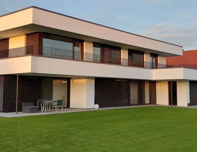 Modernes zweistöckiges Haus mit großen Fenstern, weißer Fassade und grünem Rasen vor dem Haus.