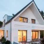 Modernes weißes Haus mit großen Fenstern, schrägem Dach und einem Balkon, umgeben von Grün in der Abenddämmerung.