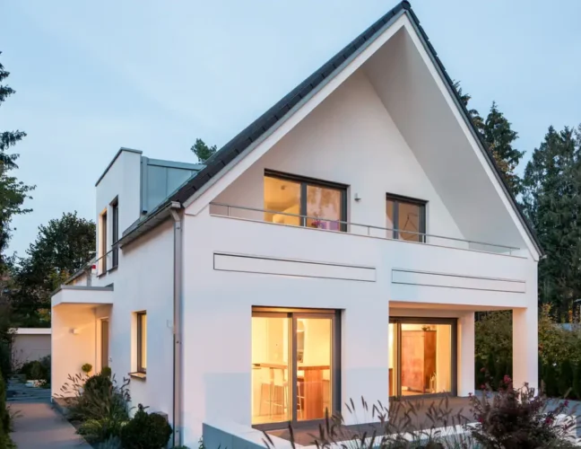 Modernes weißes Haus mit großen Fenstern, schrägem Dach und einem Balkon, umgeben von Grün in der Abenddämmerung.