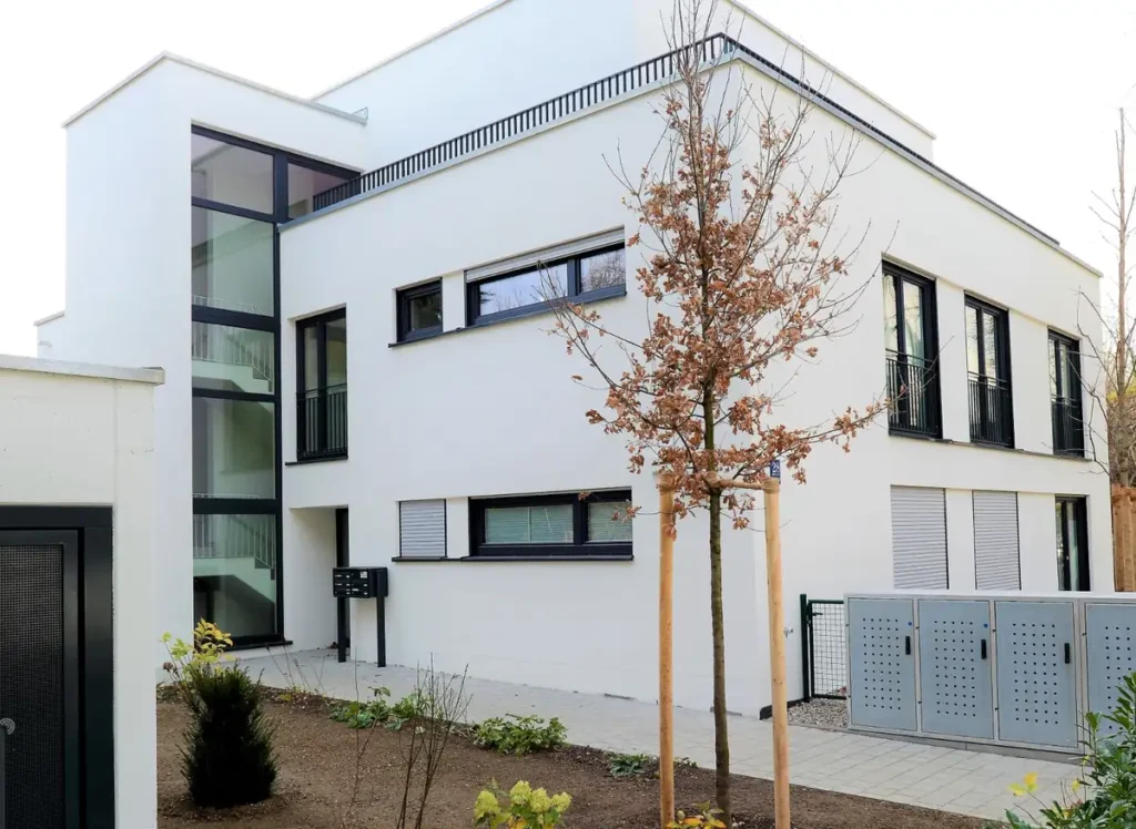 Modernes weißes zweistöckiges Gebäude mit großen Fenstern, einem kleinen Garten und jungen Bäumen vor dem Haus.