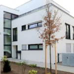 Modernes weißes zweistöckiges Gebäude mit großen Fenstern, einem kleinen Garten und jungen Bäumen vor dem Haus.