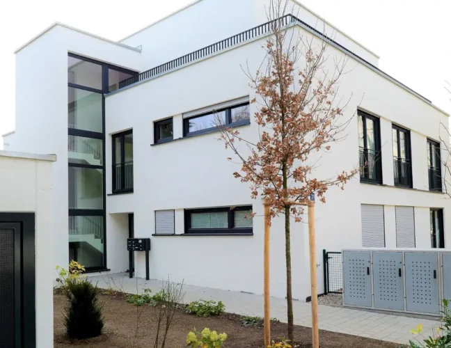 Modernes weißes zweistöckiges Gebäude mit großen Fenstern, einem kleinen Garten und jungen Bäumen vor dem Haus.
