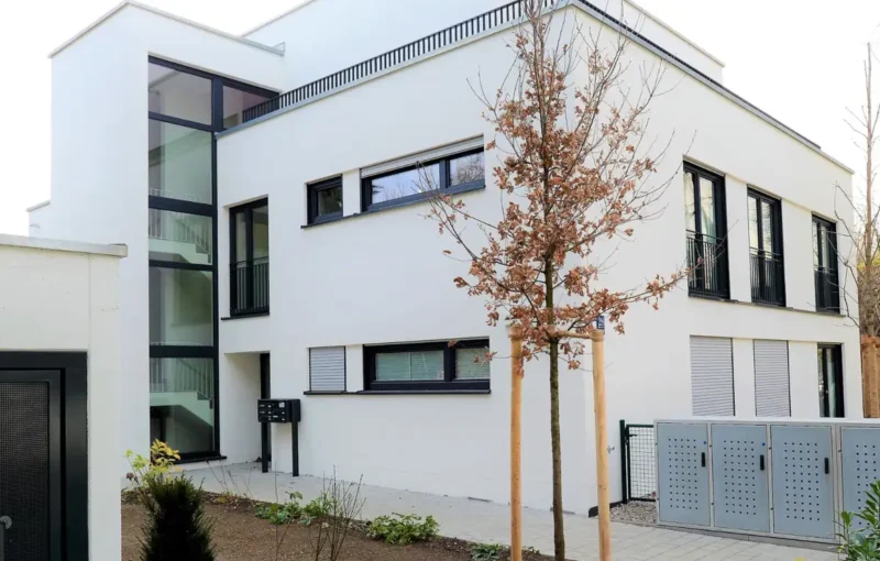 Modernes weißes zweistöckiges Gebäude mit großen Fenstern, einem kleinen Garten und jungen Bäumen vor dem Haus.