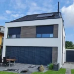 Modernes zweistöckiges Haus mit großen blauen Fensterläden, Sitzgelegenheiten im Freien und einem gepflegten Hof unter blauem Himmel.